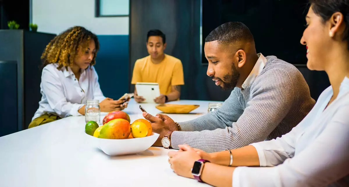 Vier Personen sitzen an einem weißen Tisch in einem modernen Büro. Zwei Personen vorne lächeln und telefonieren; eine Person trägt eine Smartwatch. Eine Frau und ein Mann hinten nutzen verschiedene Geräte. Auf dem Tisch steht eine Schale mit buntem Obst. Die Stimmung ist entspannt und kollegial.