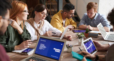 Eine Gruppe von fünf jungen Erwachsenen sitzt in einem modernen Büro um einen Holztisch und diskutiert. Laptops, Tablets und Notizbücher liegen auf dem Tisch. Die Atmosphäre wirkt kollaborativ und konzentriert, mit Pflanzen und einer Treppe im Hintergrund.