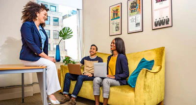 Drei Personen sitzen in einem hellen Büro. Eine Frau sitzt lächelnd an einem Schreibtisch und unterhält sich mit einem Mann und einer Frau auf einem gelben Sofa. Beide halten Laptops in den Händen. Kunstdrucke schmücken die Wände, und ein großes Fenster zeigt Gebäude draußen. Die Stimmung ist freundlich und kooperativ.