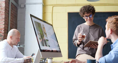 Drei Personen arbeiten in einem modernen Büro zusammen. Eine Person steht mit Tee und Tablet da und lächelt eine sitzende Person mit roten Haaren an. Eine andere Person sitzt am Computer und schaut auf ihr Handy. Auf einem großen Bildschirm läuft eine Präsentation.