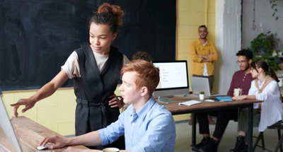 Eine Frau steht da, zeigt auf einen Computermonitor und spricht mit einem sitzenden Mann, der eine Maus bedient. Drei weitere Personen arbeiten im Hintergrund an einem Tisch zusammen, und eine Person steht lächelnd neben einer Tafel und einer gelben Wand. Die Szene ist ein modernes Büro.