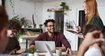 Eine Gruppe von fünf Personen sitzt und steht um einen Tisch in einem hellen, modernen Büro. Eine Person steht mit einem Tablet in der Hand und spricht, während die anderen lächelnd zuhören. Laptops, Notizbücher und Pflanzen stehen auf dem Tisch und suggerieren ein gemeinsames Meeting.