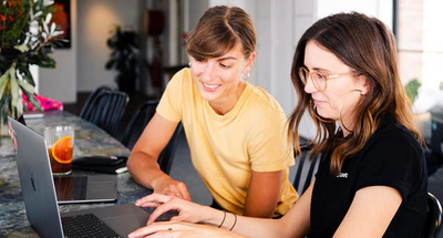Zwei Frauen sitzen an einem Tisch und arbeiten gemeinsam an Laptops. Die eine trägt ein gelbes Shirt und lächelt, während sie auf den Bildschirm der anderen blickt. Die andere, in schwarzem Shirt und mit Brille, tippt auf ihrem Laptop. Auf dem Tisch steht ein Getränk mit einer Orangenscheibe. Das Ambiente wirkt lässig und modern.